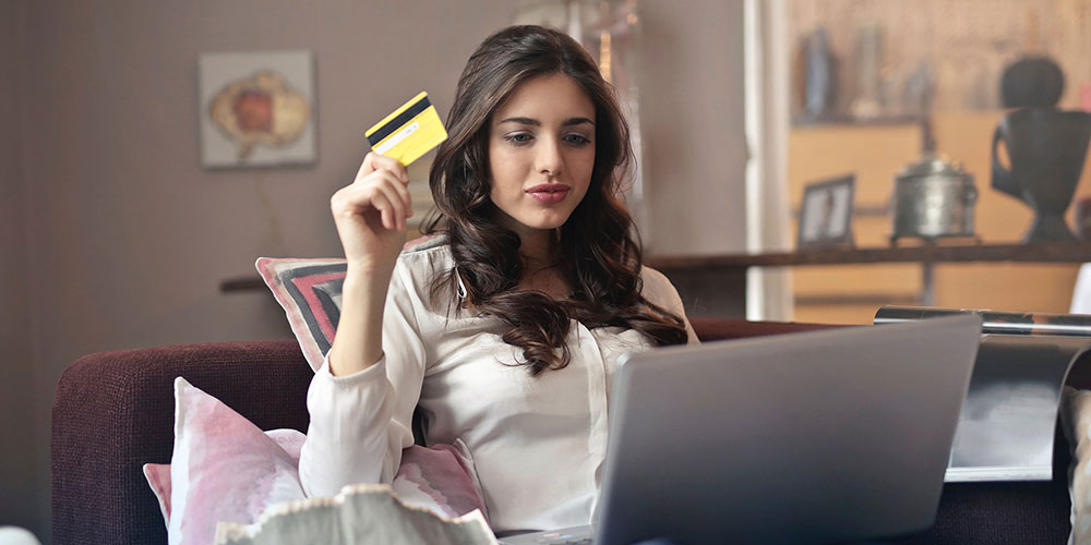 A dark-haired woman sits on a couch with a laptop open and a yellow credit card in her hand.