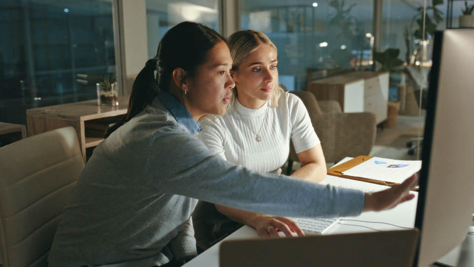 Two women sitting at a desk looking at the same screen. One of them is point at the screen.