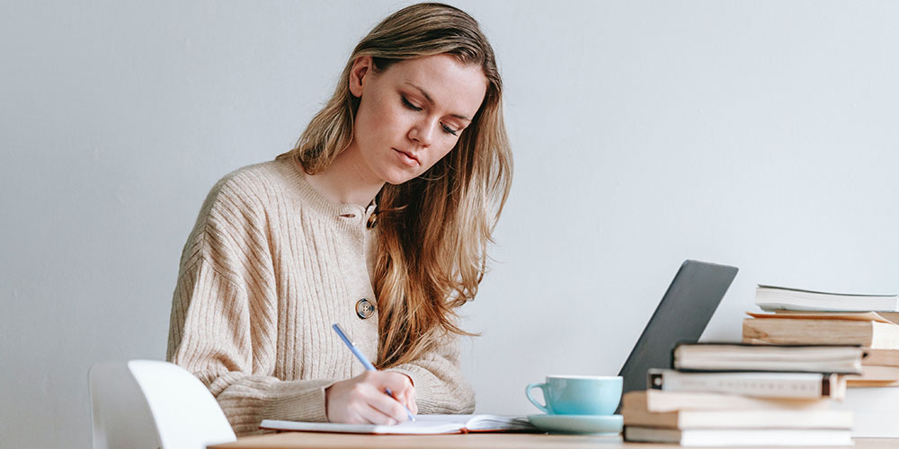 a young woman writes in her journal. A laptop, tea cup and stack of books are on the table in front of her.