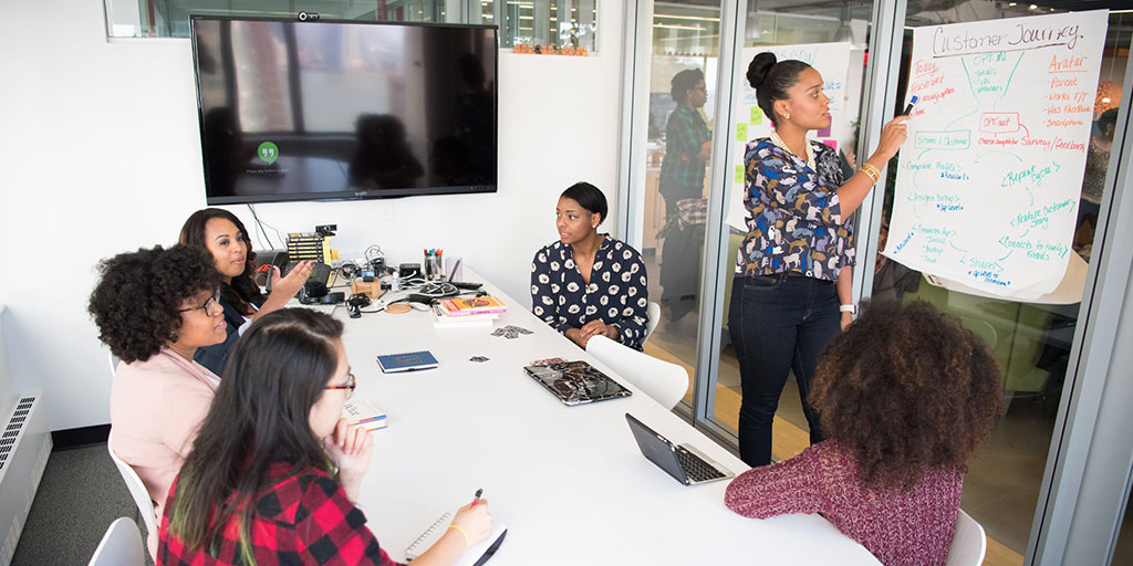 A group of people sit around a table in a meeting room. A women is standing and writing on a whiteboard