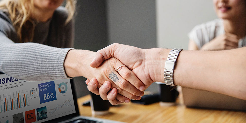 A man wearing a watch with a silver band shakes hands with a woman wearing a silver ring. She has a tattoo on her middle finger.