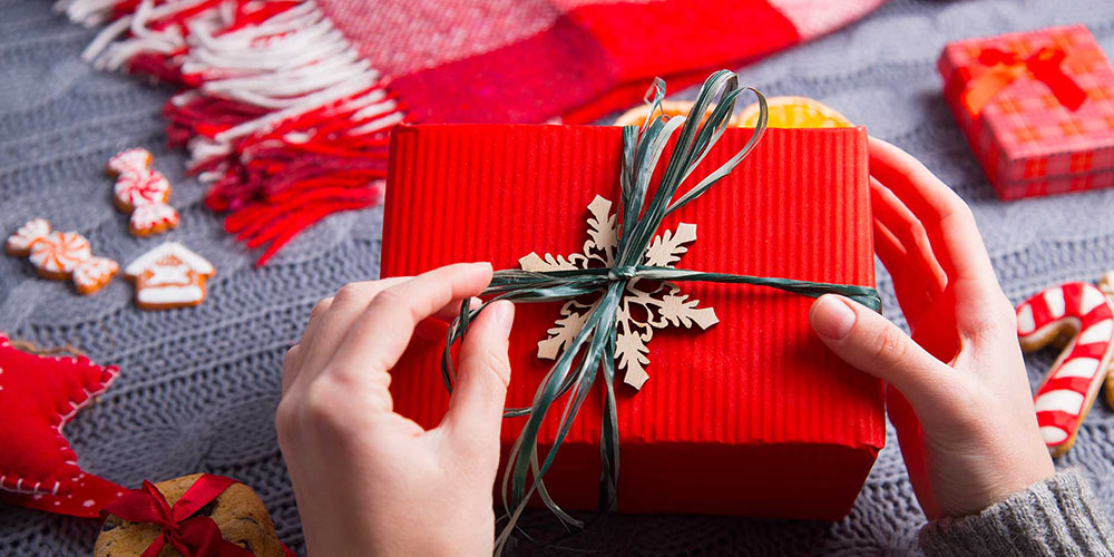 A woman’s hands are seen wrapping a box in red paper with a cardboard snowflake and green raffia to tie it.