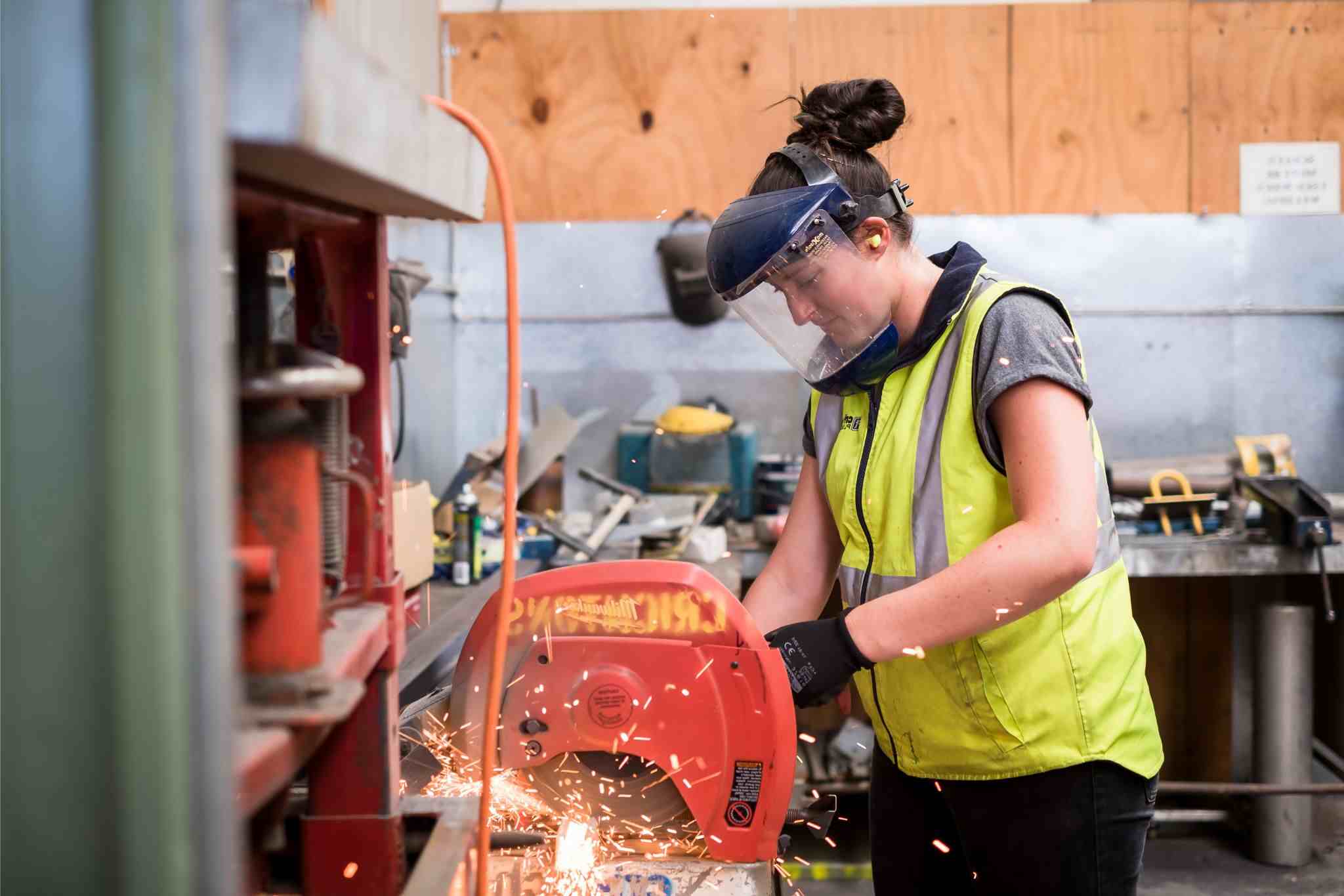 A woman at a workbench wearing protective equipment. She is working with a powered saw to cut materials.
