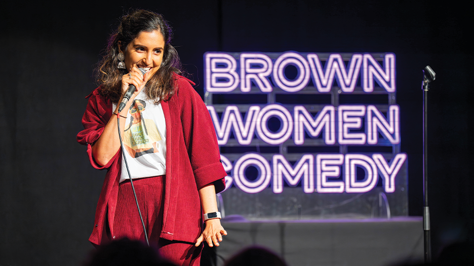 Daizy Mann on stage in front of a sign reading 'Brown Women Comedy'.