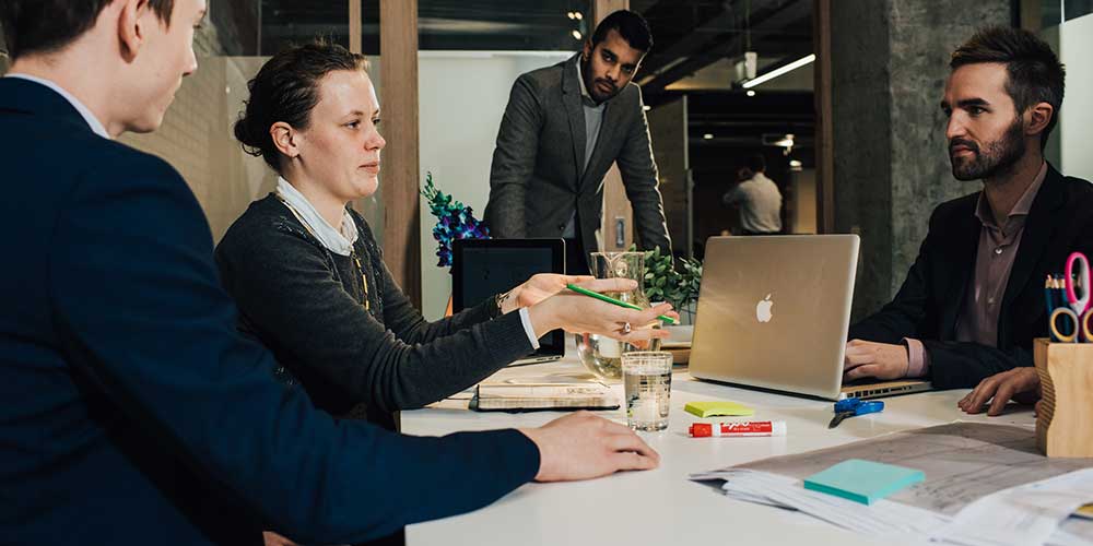 A woman and two men sit at a meeting table. A bearded man stands at the end of the table looking on.