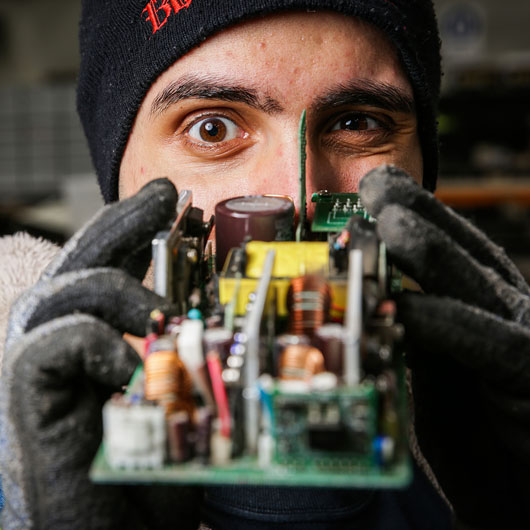 A man holding a circuit board. He has his face behind the board looking at it closely.