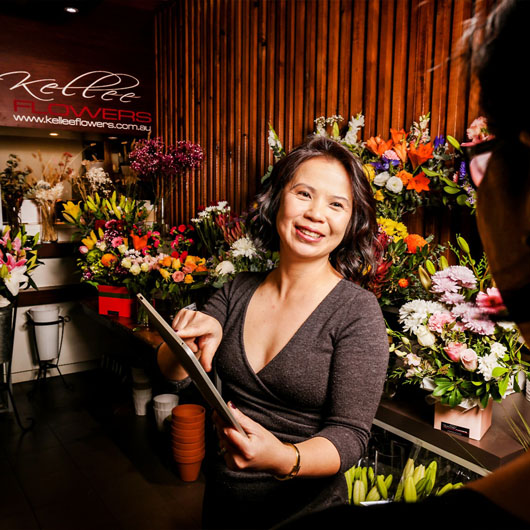 A woman holding a clipboard in a flower shop. Another person is looking on.