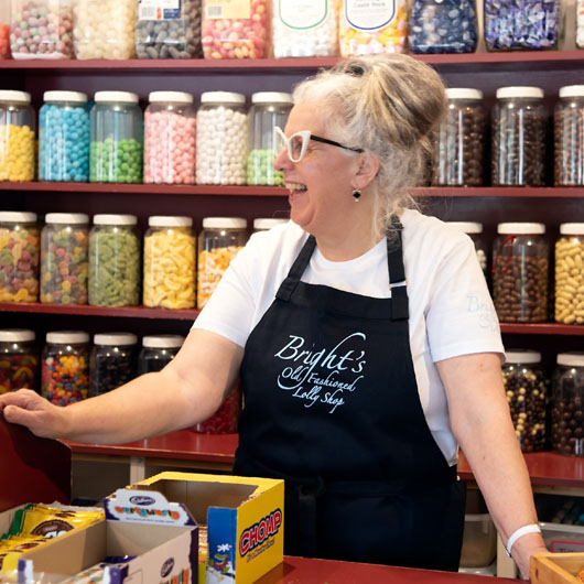 A woman working at Brights Old Fashioned Lolly Store laughing and looking off camera. She is behind the store's bench. On the wall are jars of different lollies.