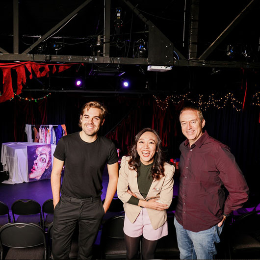 Three people standing in a small theatre venue smiling at the camera. The stage is behind them.