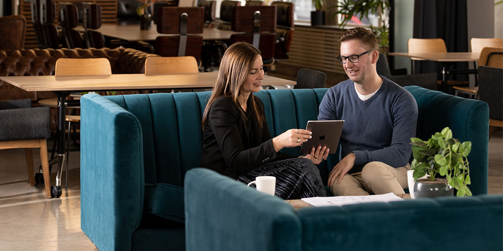 A well dressed man and a woman sit on a teal couch in a co-working space looking at an ipad held by the woman. 