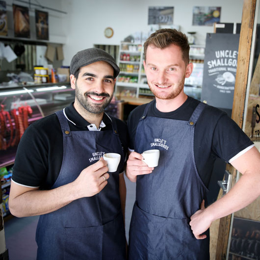 Two men working at Uncle's Smallgoods butcher. They are holding small coffee cups, smiling at the camera.